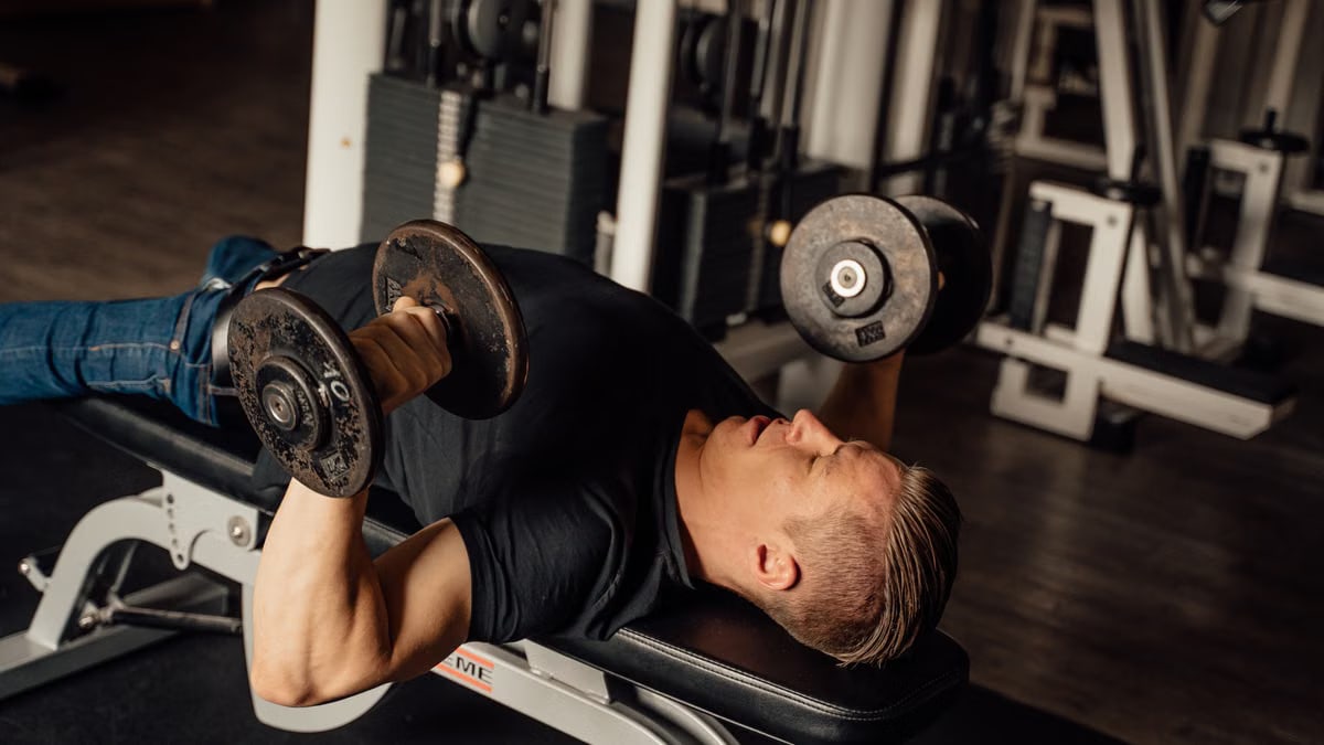 A man exercises with dumbbells on a bench press at a gym, focusing on fitness and strength.